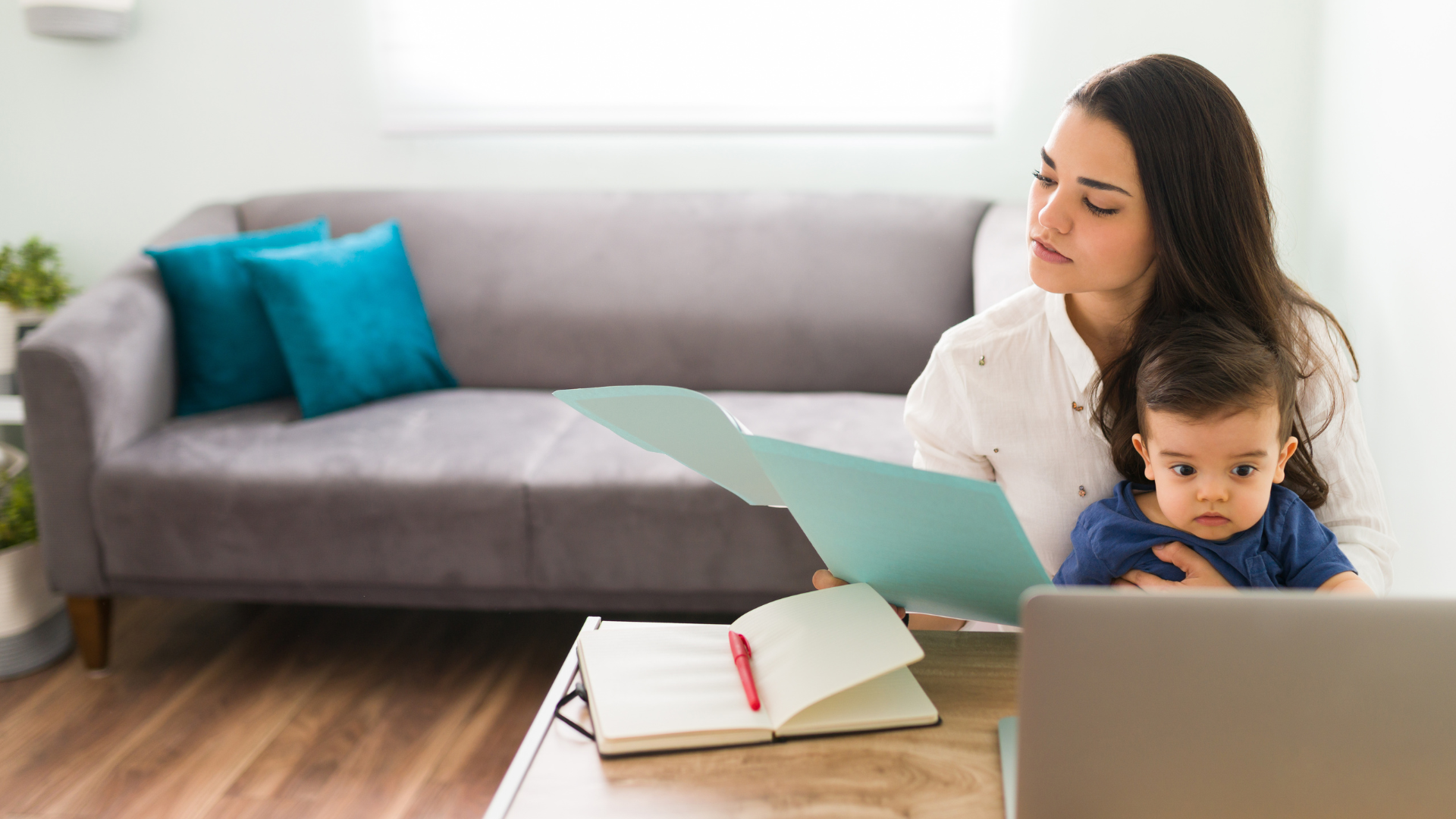 Woman working with laptop and notebook in her living room with a baby on her lap