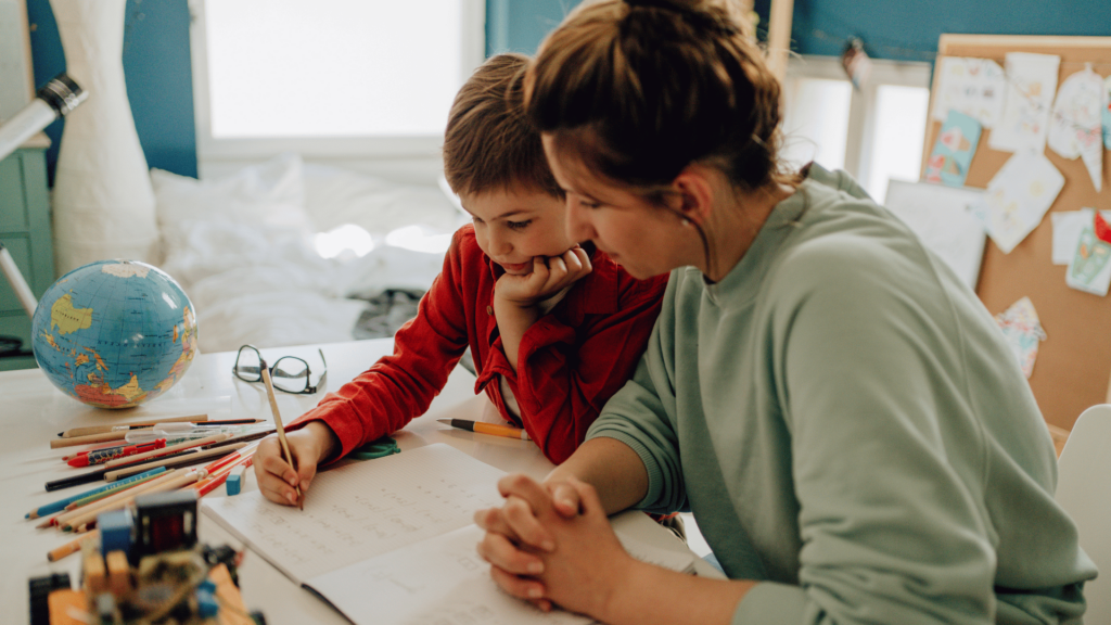 Mom sits next to her son at a desk, helping him practice a writing assignment.