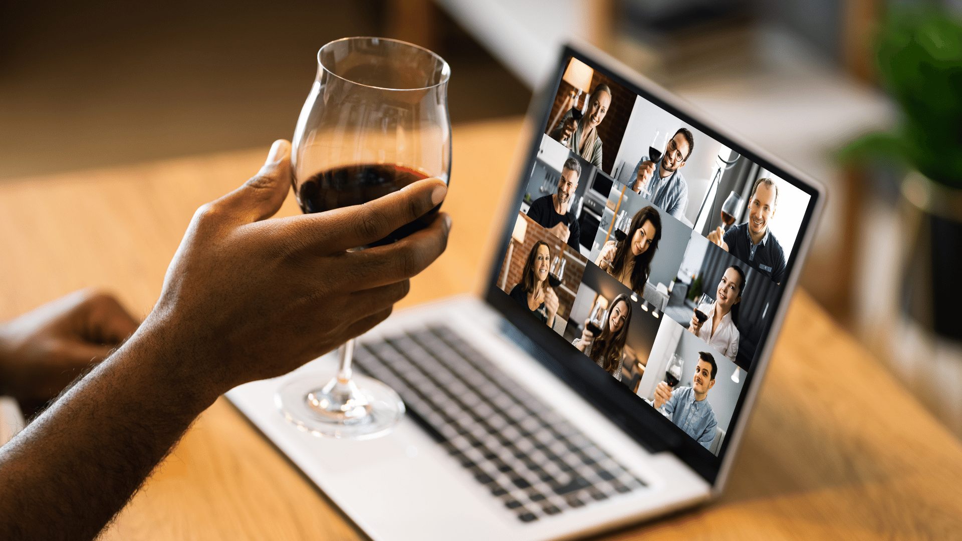 Image of a laptop open with a video conference call with several people, and a person's hand in the foreground holding a glass of wine.