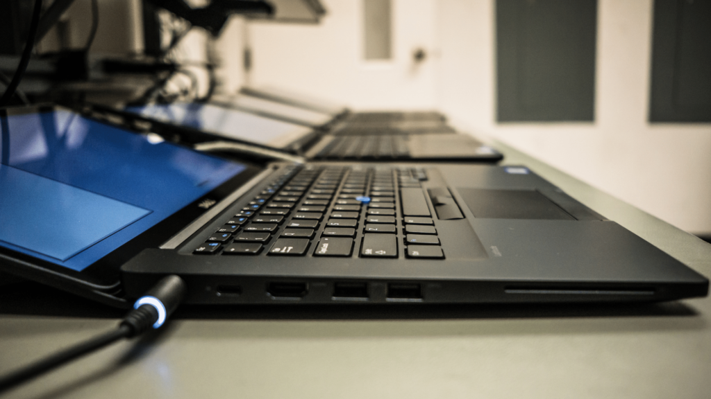 Close-up photo of a laptop, open and charging, with several similar laptops in the background.