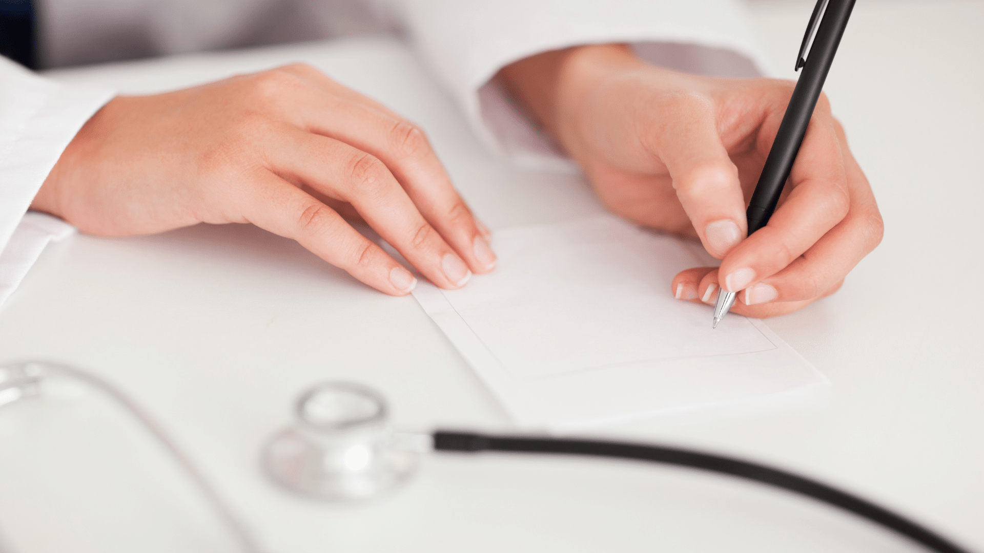 Close up of a doctor's hands holding a pen and notepad with a stethoscope in the foreground.