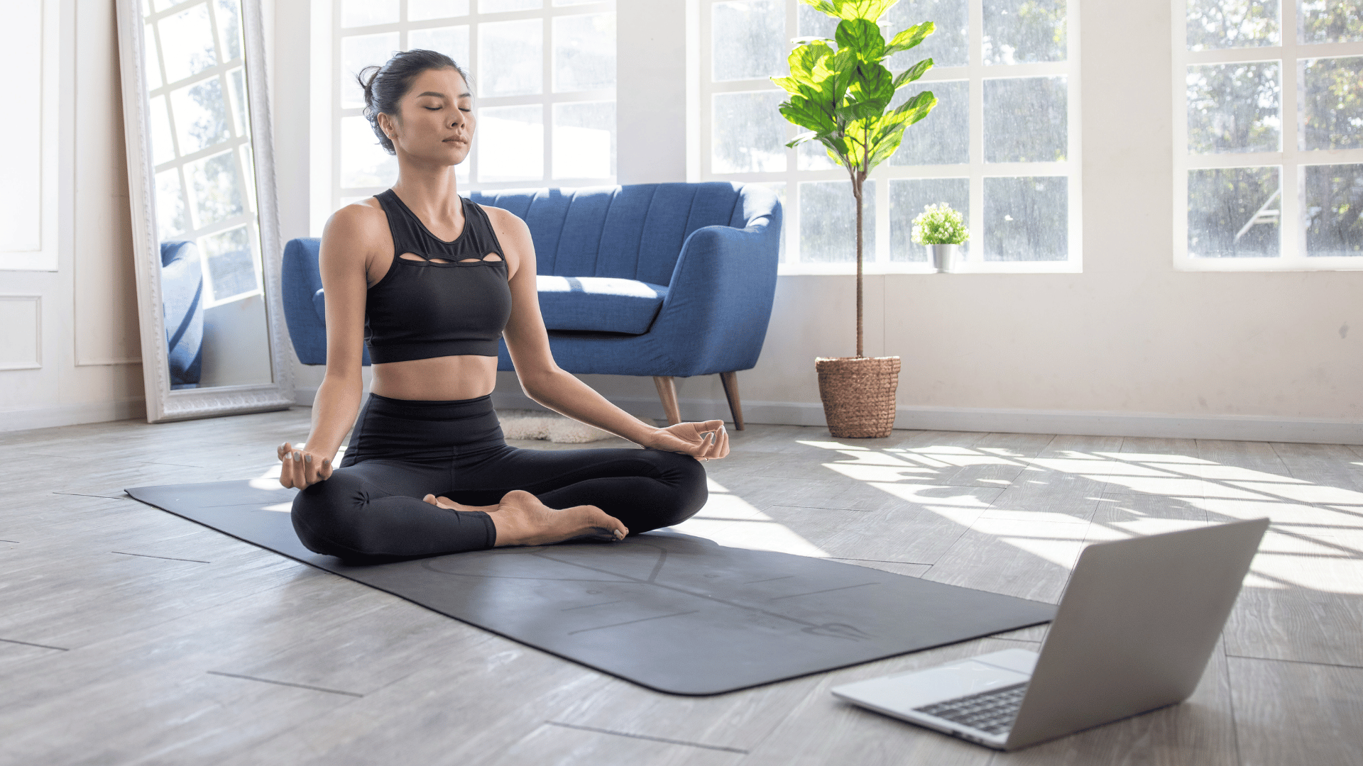 Woman sitting cross-legged on yoga mat at home, with a laptop open in front of her.